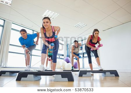 Full length of instructor with fitness class performing step aerobics exercise with dumbbells in a gym
