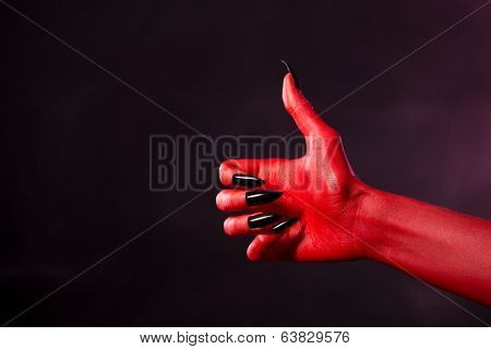 Spooky red devil hand with black nails showing thumbs up, studio shot on smoky background 