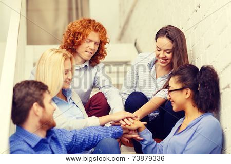 business, office, gesture and startup concept - smiling creative team with hands on top of each other sitting on staircase