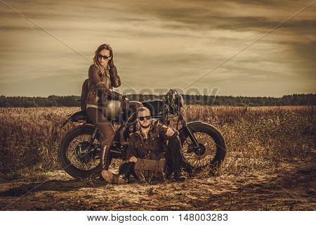 Young, stylish cafe racer couple on the vintage custom motorcycles in a field.