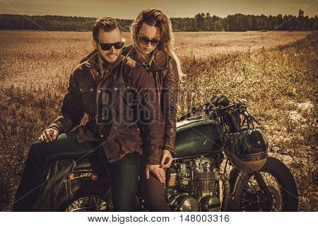Young, stylish cafe racer couple on the vintage custom motorcycles in a field.