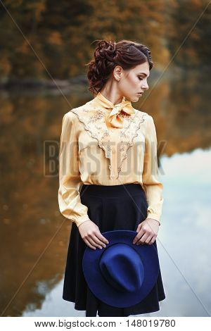 portrait of young brunette woman with hairstyle in elegant vintage blouse with bow and skirt with fashionable hat in hands near the river or lake in autumn park. Woman with hat in autumn park. Young beautiful model posing near the water.