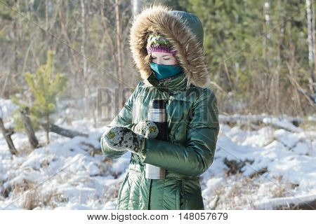 Concentrated girl is wearing green shiny down coat opening stainless steel thermos flask in winter frosty wood outdoors
