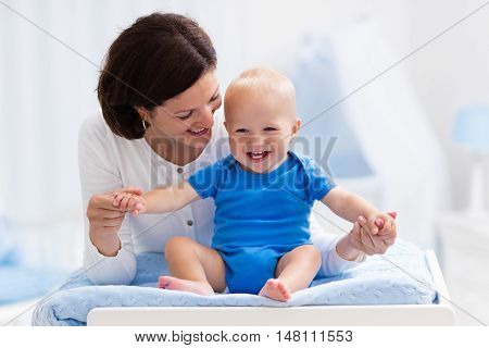 Young mother taking care of little boy in white sunny nursery with changing table baby crib and rocking chair. Diaper change and clothing. Mom and son in beautiful bedroom at home.