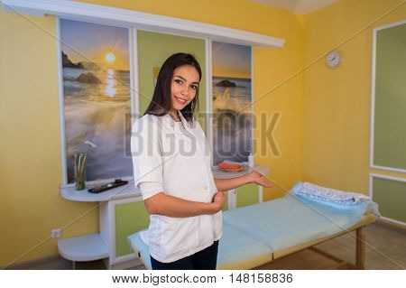 Smiling beauty therapist standing beside massage table at the spa and welcomes invites customer.
