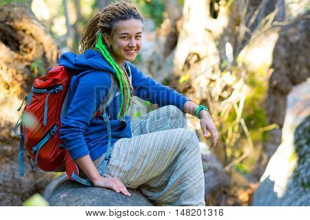 Cute smiling Girl with Day Backpack and sporty casual Clothing sitting on large fallen Tree Stalk in wild Forest Outdoors Sunlight