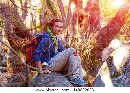 Cute smiling Girl with Day Backpack and sporty casual Clothing sitting relaxed on large fallen Tree Stalk in wild Forest Outdoors enjoying warm Sunlight
