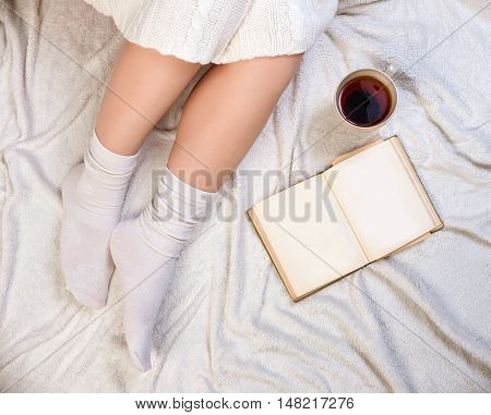 Long female feet in warm woolen socks around her feet is a book she is reading it. Part of the body. Beautiful female legs sitting on the bed with a cup. Morning with a book