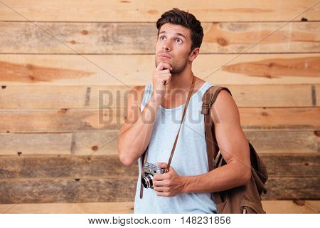 Portrait of a pensive photographer with backpack holding camera isolated on a wooden background