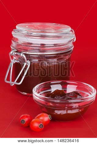 Rose hip jam and fruits over red background, also rose haw or rose hep. Marmelade in glass jar and bowl. Shells of ripe fruits can be eaten raw and are one of the richest vitamin C sources in plants.
