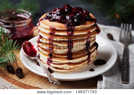 Stack of american pancakes or fritters with strawberry and blueberry jam in white plate on wooden rustic table decorated Christmas tree, vintage style.