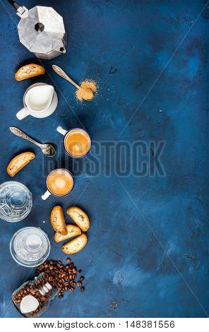 Coffee espresso in cups with italian cantucci, cookies and milk in jug over dark blue painted plywood background, top view, copy space. Food frame concept