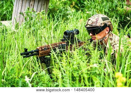 Military man sniper with automatic rifle with a telescopic sight lies in grass in forest. Airsoft