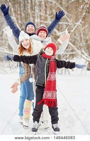 Smiling family of four stands on skates on ice pathway in winter park spreading arms to sides.