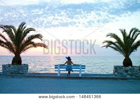 Lonely young girl in a hat sitting on a bench on the beach with palm trees at sunset