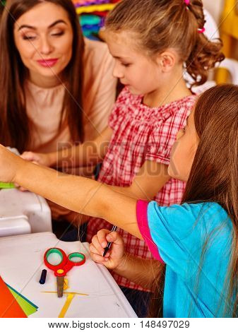 Close up of children girl with young female teacher are making something out of colored paper on table in primary school. Children craft lesson in primary school. Teacher learn children.