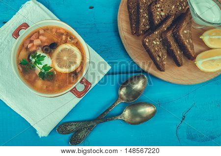 Solyanka - Russian traditional meat soup with hot spices in a bowl with bread and spoons on blue wooden background. Meat solyanka - traditional Russian dish. Russian cuisine concept. Top view.