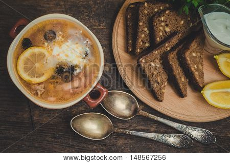 Solyanka - Russian traditional meat soup with hot spices in a bowl with bread and spoons on old wooden background. Meat solyanka - traditional Russian dish. Russian cuisine concept. Top view.