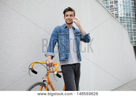 Smiling guy in denim jacket with beard and orange bicycle standing on the background of white wall. Student calling on the phone