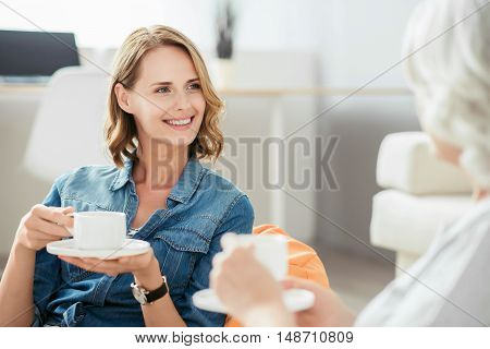 Nice time spending. Cheerful charming smiling woman drinking coffee and talking with her mother while resting at home