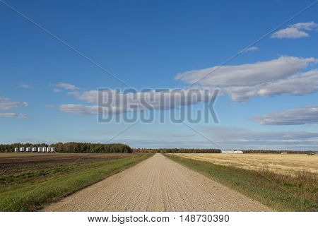 Gravel road dividing harvested fields with grain storage bins and equipment and autumn forest in the distance