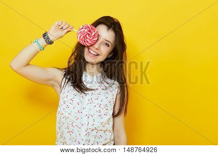 happy modern teenage woman smiling and hiding behind a lollipop candy over yellow background