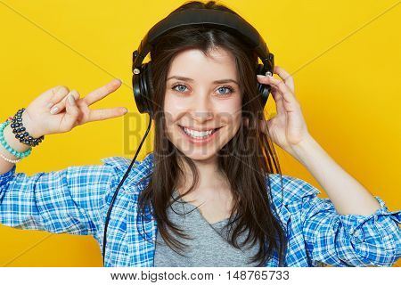 Young woman wearing headphones over bright yellow background. Hispter girl listen to music and showing peace sign