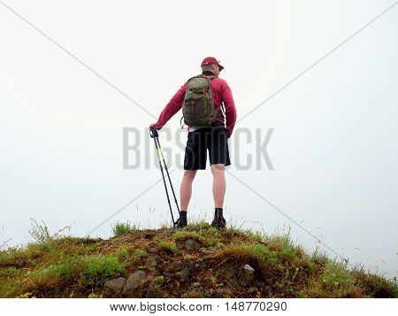 Man in cloud of fog. Hiker in pink black sportswear and poles stand on mountain peak rock. Around heavy mist. Poor visibility