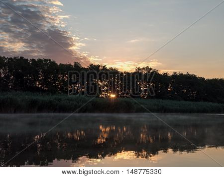 Sunrise in the river with reflex. Mirrored forest line on the river. Fantastic foggy river with fresh green grass in the sunny beams. Dramatic colorful scenery.
