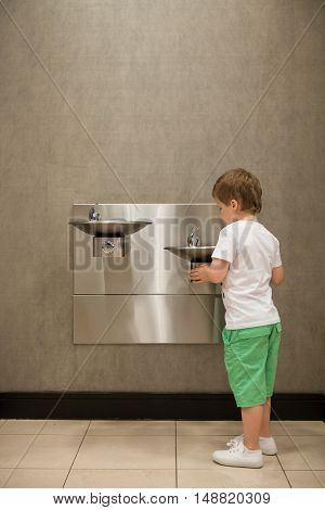 Curious kid boy drinking water from drinking fountain. Stainless Steel drinking fountains mounted at different heights. Young customer in the shop.