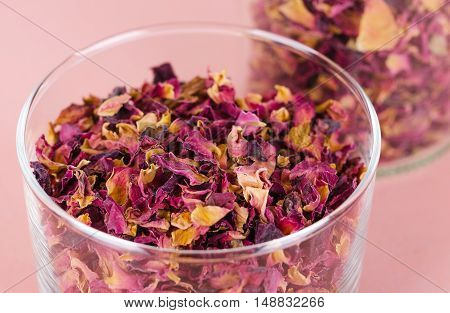 Rose petals in a glass bowl on rosy background. Dried blossoms, used for perfumes, cosmetics, teas and baths. Purple and orange colored organic herb. Isolated macro photo close up from above.