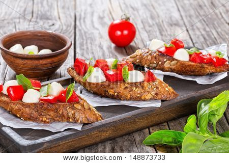 bruschetta with caprese salad on rye baguette with seeds on parchment paper on dark wooden board. and on clay dish italian recipe close-up view from above