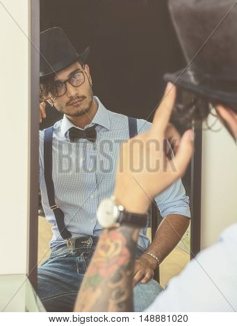 Portrait of brunette young man in glasses, hat, bow-tie, suspenders and shirt looking at himself in a mirror. Vintage look