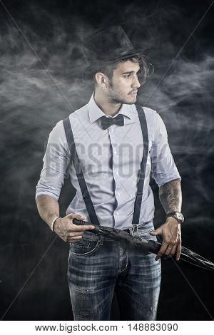 Portrait of brunette young man in glasses, hat, bow-tie, suspenders and shirt on dark smoky background. Vintage look