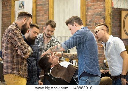 Teacher training group of students in beard shaving with metal straight razor in hairsalon. Interior shot of learning process in modern barbershop