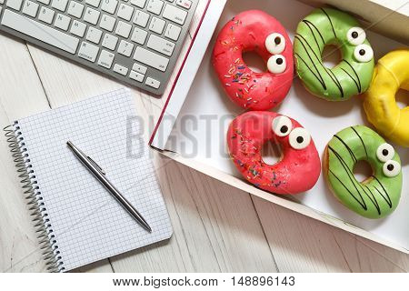 Box of colorful glazed donuts with funny eyes on the office desk near computer keyboard and notepad with pen. White rustic wooden table with business lunch snack dessert top view