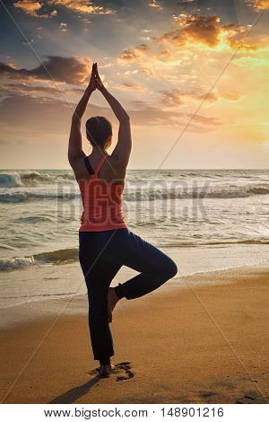 Yoga outdoors - sporty fit woman doing Hatha yoga asana Vrikshasana tre pose on tropical beach on sunset