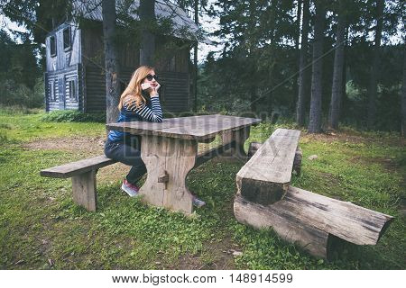 Young woman sitting at a table near the old wooden house in the forest