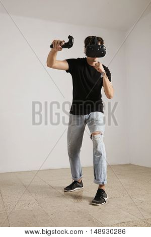 Full body shot of a young gamer in VR glasses and jeans and black unlabeled t-shirt playing a game in a room with white walls and light wooden floor.