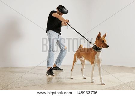 A young gamer in VR headset and black unlabeled t-shirt pulling a leash on a brown and white basenji dog against a white wall background.