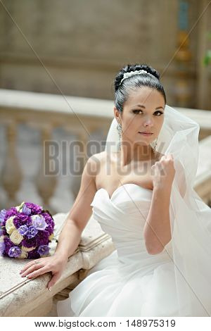 Beauty bride in bridal gown with bouquet and lace veil indoors. Beautiful model girl in a white wedding dress. Female portrait of cute lady. Woman with hairstyle