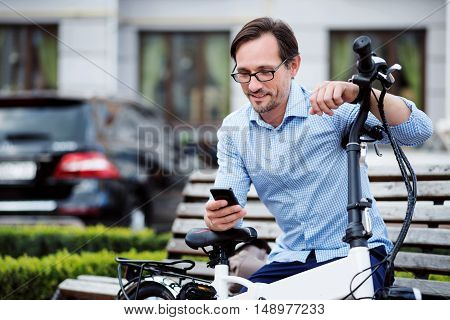 Virtual communication. Handsome bespectacled man putting his hands on bike and using laptop while sitting on the bench.