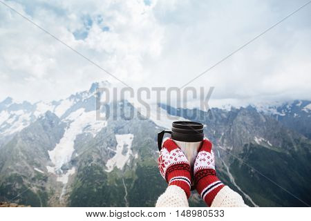 Closeup photo of thermos mug with tea in traveler's hand over out of focus mountains view with snow, tourizm in cold season