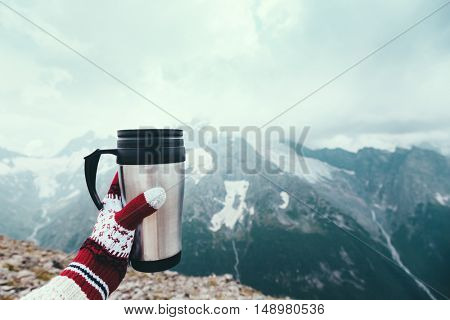 Closeup photo of thermos mug with tea in traveler's hand over out of focus mountains view with snow, tourizm in cold season