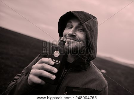 VIK, ICELAND - MAY 04, 2018: Hooded man holding a bottle of Valhalla, a herbal liqueur from the Finnish Koskenkorva. Icelandic landscape in the background