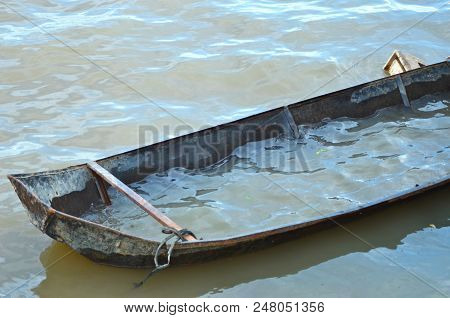An Old Tin Boat Is Half-filled With Water And Moored On A Muddy River. The Scene Is Deserted.