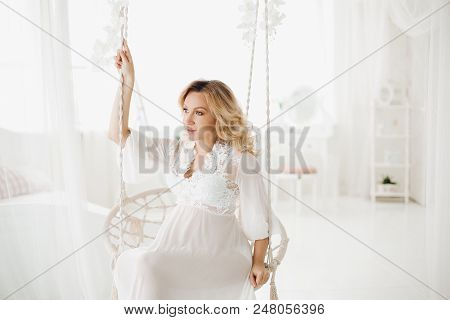 Beautiful Blonde Pregnant Woman Wearing In White Long Dress Sitting At Swing With Flowers And Lookin