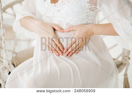 Beautiful Blonde Pregnant Woman Wearing In White Long Dress Sitting At Swing With Flowers And Lookin