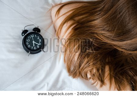 Young Woman Sleep With Alarm Clock On The Bed In The Morning. Unrecognizable Girl With Long Hair And