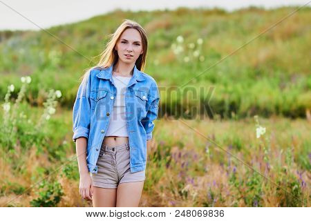Happy Smiling Young Girl Wearing Blue Jeans Jacket And Shorts Is Standing In The Field
